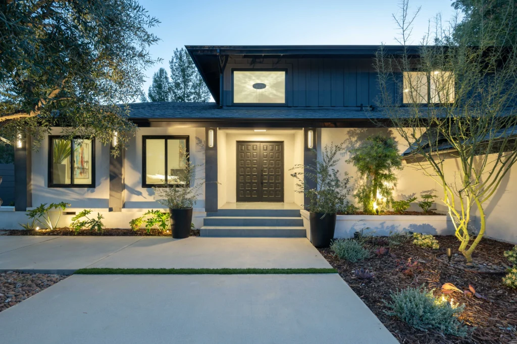 A slightly elevated view of the main entrance to a contemporary house. Steps lead up to a dark modern wood front door flanked by large dark-framed windows. Spotlights and path lights illuminate the scene, emphasizing the dark wood second-story cladding and the low-water landscaping.