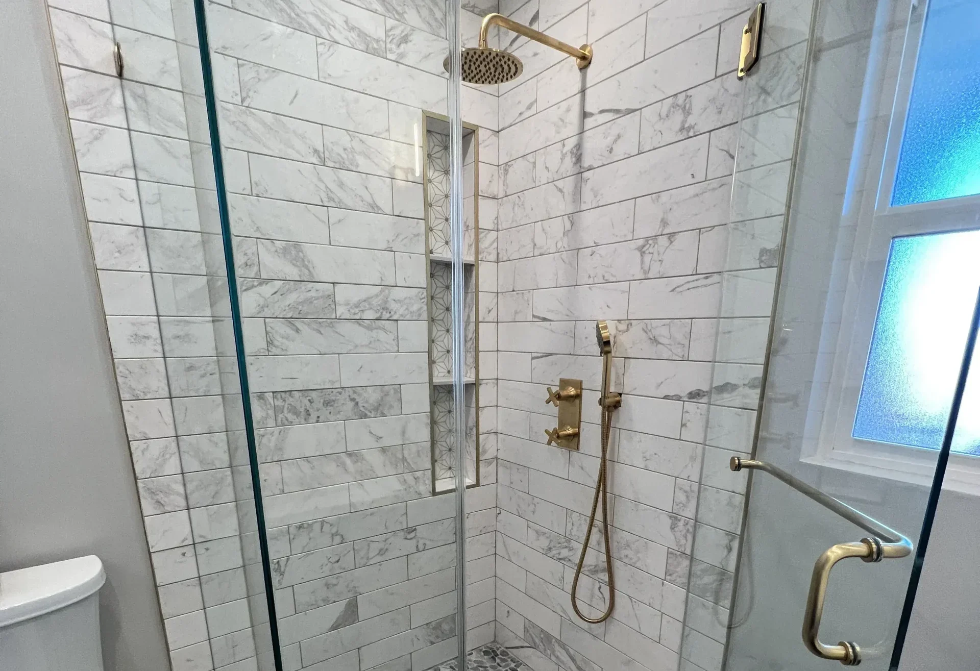 A view of the shower entrance with a gold handle, showing the contrast between the pebble floor and the bathroom's wood-look tiling.
