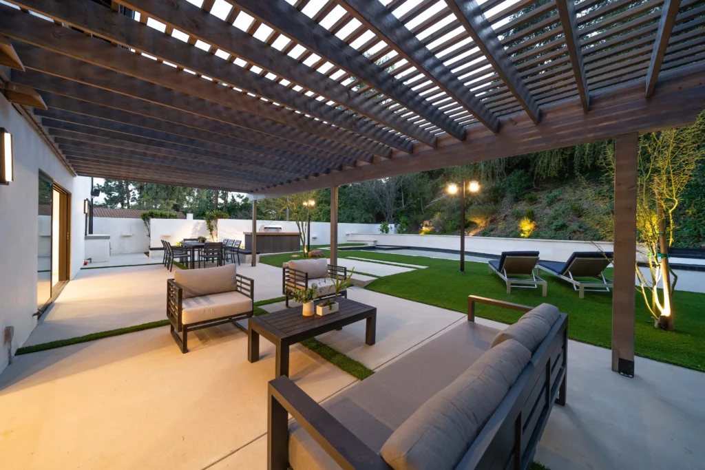 An outdoor shot of a modern backyard patio with grey concrete pavers and synthetic grass. A large dark wood pergola with lattice roofing covers a complete lounge set (sofa and chairs) and a dining table with chairs. String lights hang above.