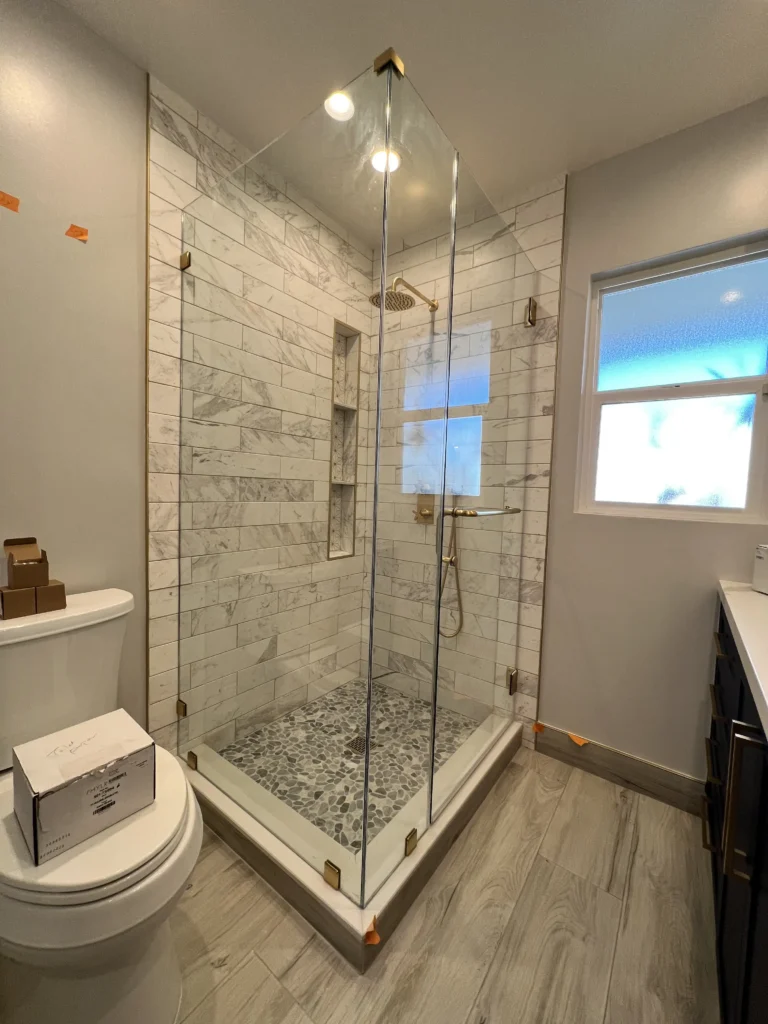 A corner view of a bathroom featuring a frameless glass shower with marble-patterned wall tiles and a pebble floor, positioned next to a white toilet.