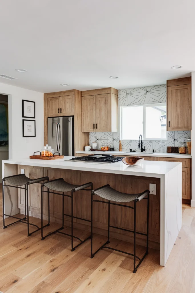 A vertically oriented photograph of a kitchen island with light-wood cladding and a waterfall-edge white quartz countertop. Three backless, grey woven-seat barstools are positioned underneath.