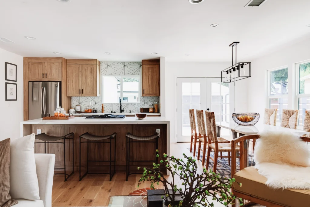 A medium-wide shot of an open-plan living and dining space. The kitchen island and cabinetry are visible to the far left. The focus is on the dining area with a wooden table, woven chairs, and a black chandelier.