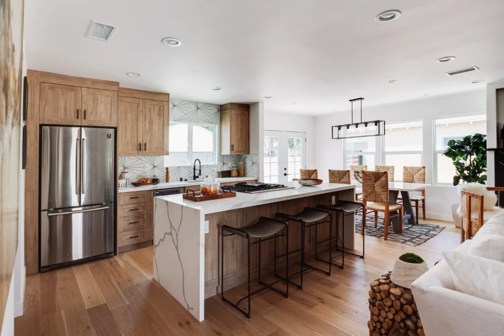 A wide-angle landscape photograph capturing an entire open-plan great room, merging a modern kitchen (left), a central dining area, and a living space (right). The kitchen has light-wood cabinets, a large island with a waterfall counter and grey stools.