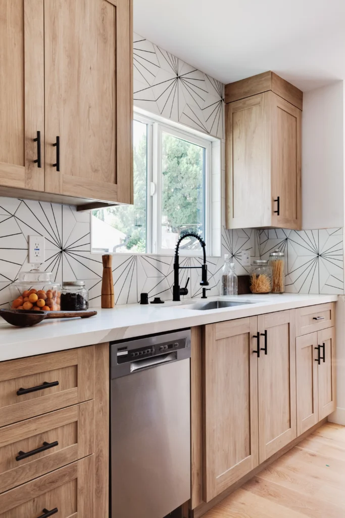 A vertical shot of a kitchen featuring light oak cabinets with black handles, a white quartz countertop, and a black gooseneck faucet. The backsplash consists of white tiles with a black starburst geometric pattern.