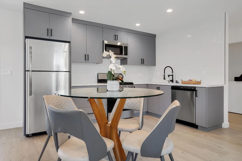 A professional interior photo of a grey-paneled shaker kitchen. From the left, a stainless steel refrigerator is visible, followed by lower cabinets, a grey range oven, upper cabinets, a dark backsplash, a black gooseneck faucet over a sink, and a view into a living area.