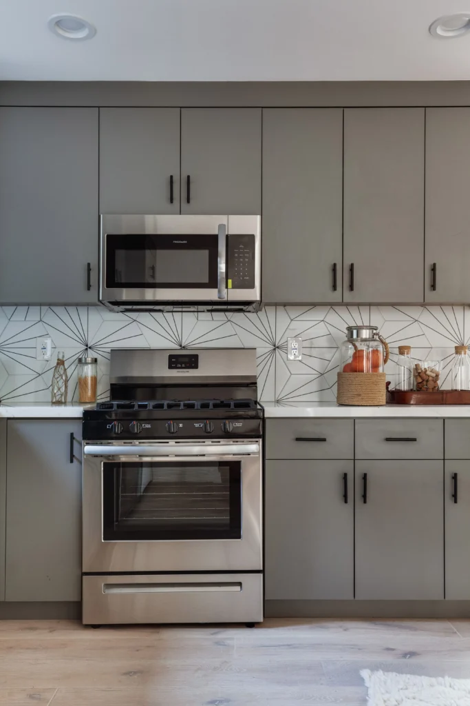 A straight-on, symmetrical shot of a stainless steel gas range and microwave surrounded by matte grey cabinets and a geometric tile backsplash.