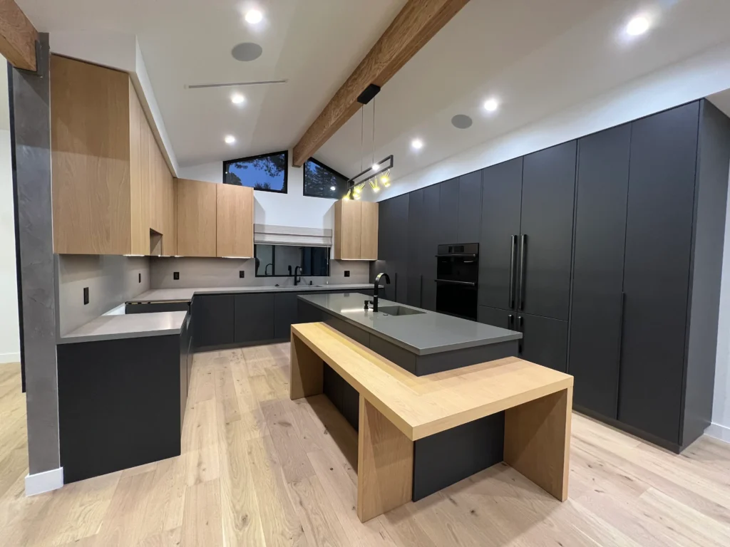 A modern kitchen featuring matte black floor-to-ceiling cabinetry, light wood upper cabinets, a grey stone island with an attached wood breakfast bar, and vaulted ceilings with exposed timber beams.