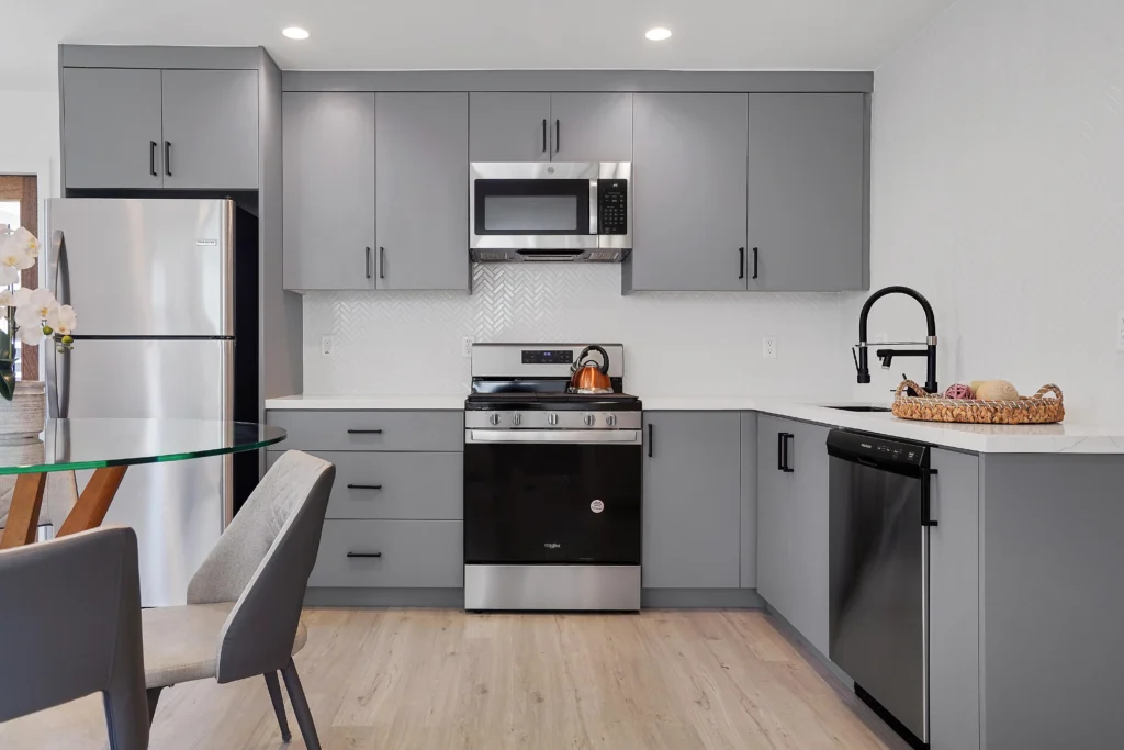 A professional interior photo of a grey-paneled shaker kitchen. From the left, a stainless steel refrigerator is visible, followed by lower cabinets, a grey range oven, upper cabinets, a dark backsplash, a black gooseneck faucet over a sink, and a view into a living area. In the blurred foreground, a glass dining table edge and chairs are seen. The floor is light wood.