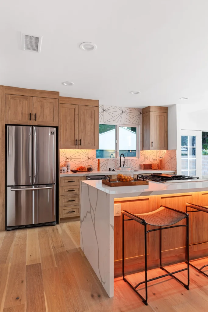 A wide shot of the oak kitchen with warm orange LED under-cabinet and under-island lighting turned on, highlighting the wood grain and waterfall counter edge.