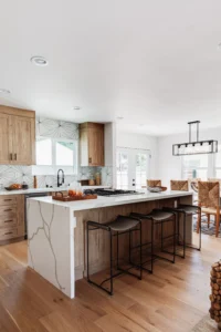 A wide-angle view of an open-concept kitchen and dining area. A large white quartz waterfall-edge island with three woven stools sits in the foreground. A dining table with woven chairs is visible in the background.