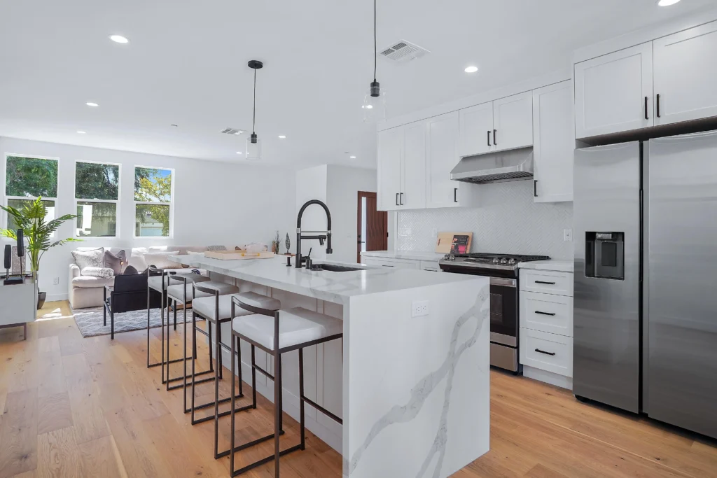 A wide-angle interior photograph capturing the expansive modern kitchen and living space. A large white kitchen island with a marble-patterned waterfall edge dominates the center-left, with two modern stools.