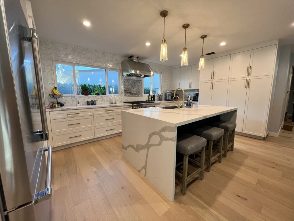 A wide view of a modern kitchen with white cabinetry, a waterfall island with gray veining, and warm wood flooring.