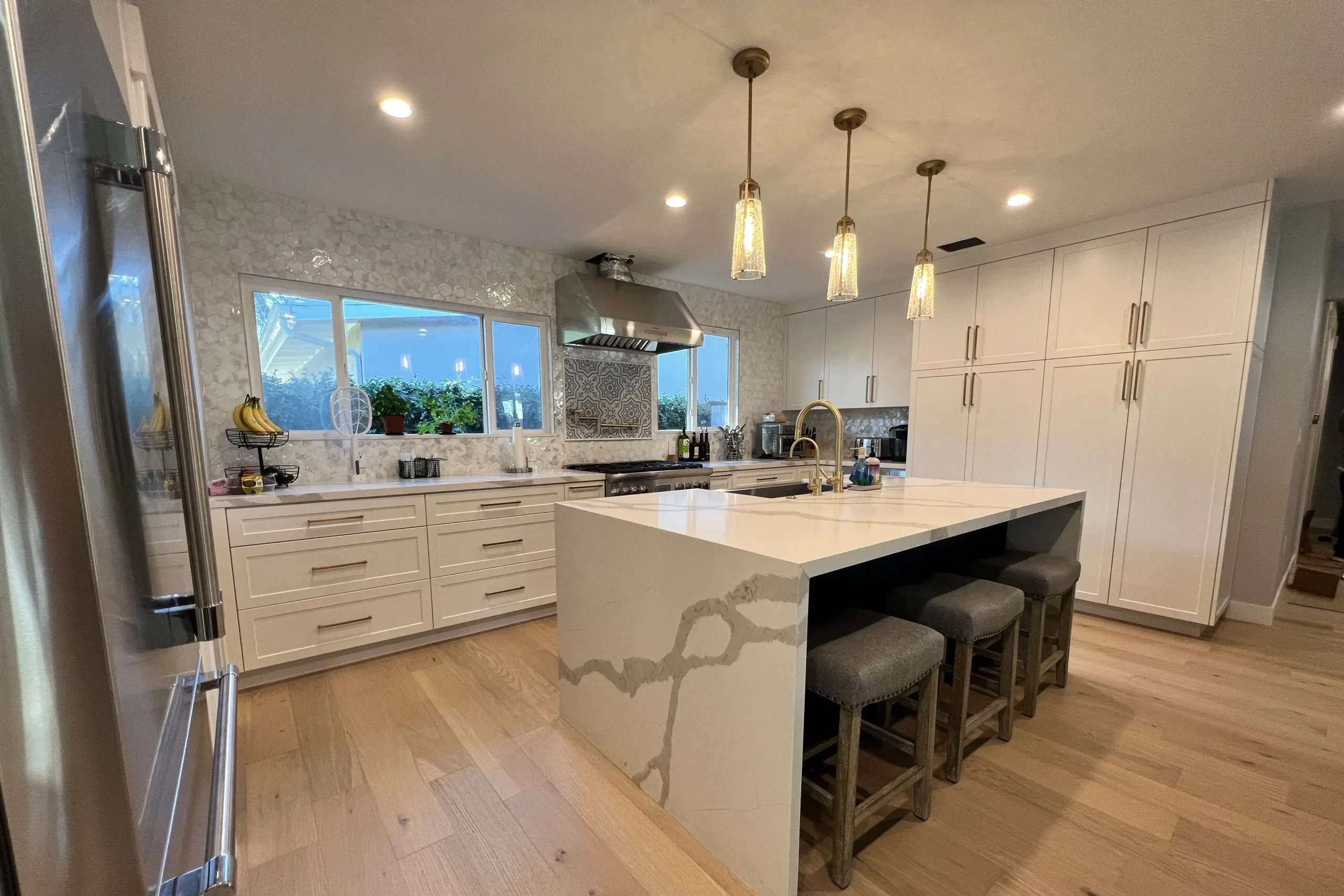 A wide view of a modern kitchen with white cabinetry, a waterfall island with gray veining, and warm wood flooring.