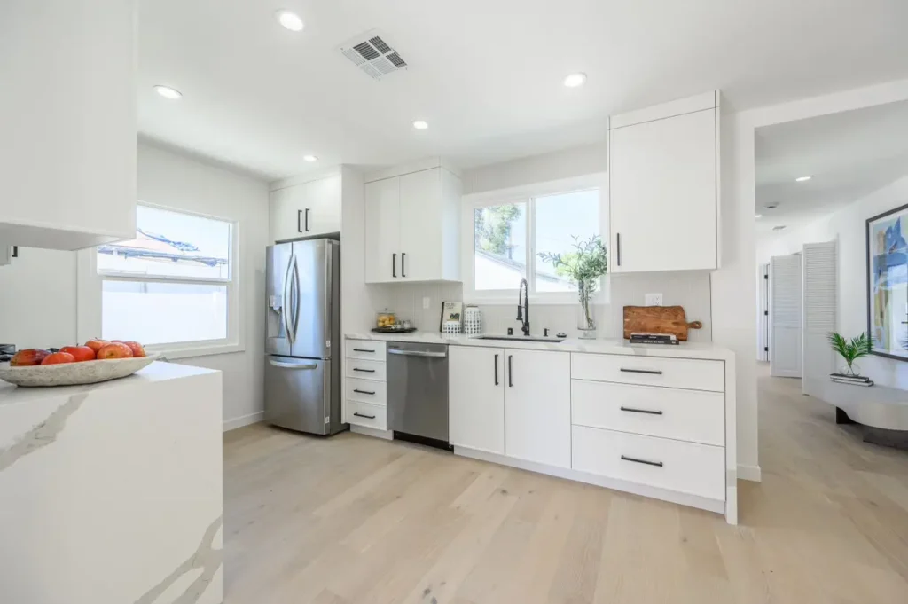 A wide view of a modern white kitchen with light wood floors and stainless steel appliances.