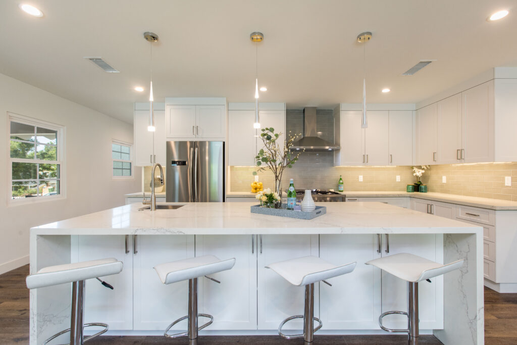 Modern white kitchen featuring a large marble-topped island with breakfast bar seating and stainless steel appliances.