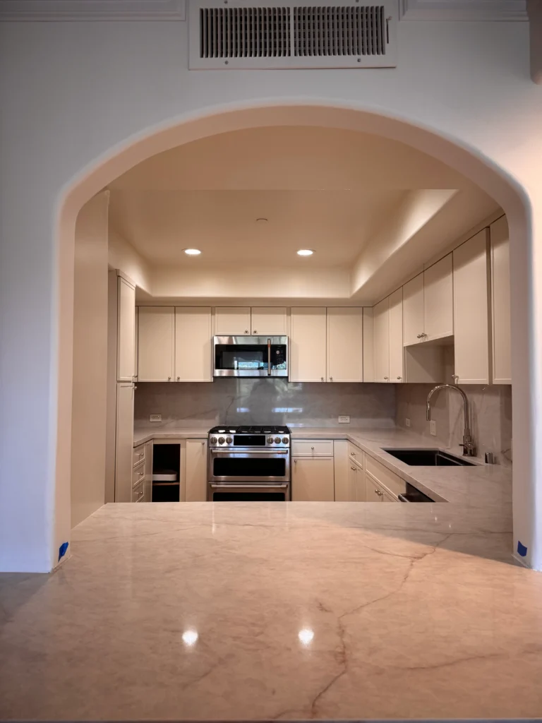 A bright kitchen with white cabinets seen through a white archway under warm recessed lighting.
