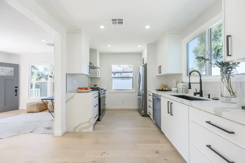 Looking through a kitchen with white cabinets and black pulls toward a bright dining or living area.