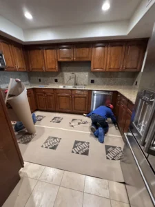 View of a kitchen floor covered in protective cardboard with workers installing or measuring cabinets.