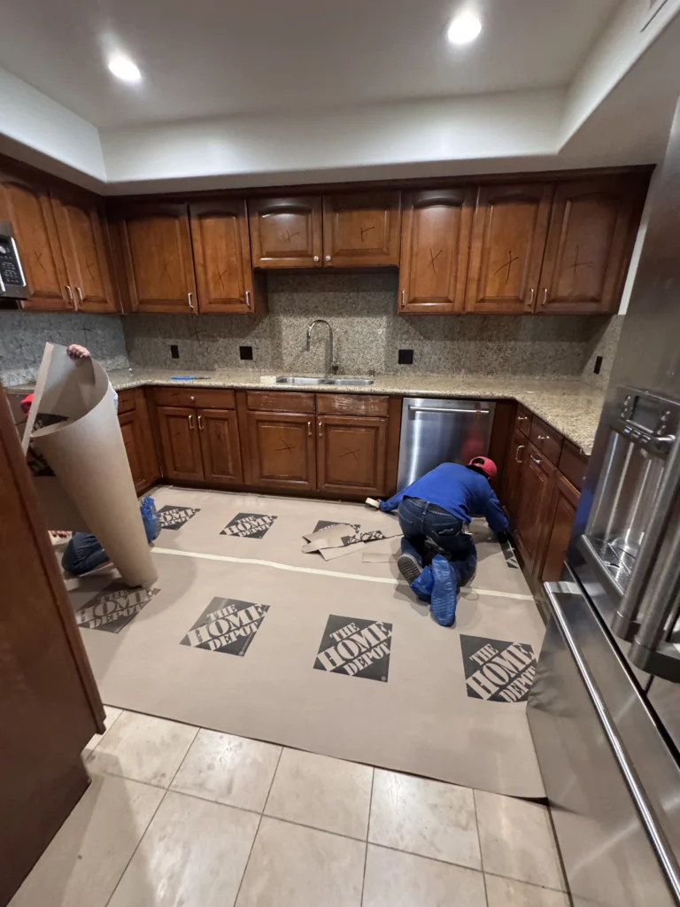 View of a kitchen floor covered in protective cardboard with workers installing or measuring cabinets.