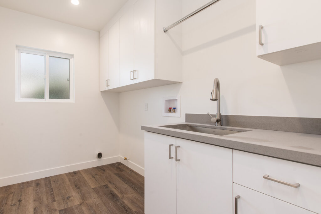 A clean, white laundry room with grey quartz countertops, a deep stainless steel utility sink, and ample cabinetry.