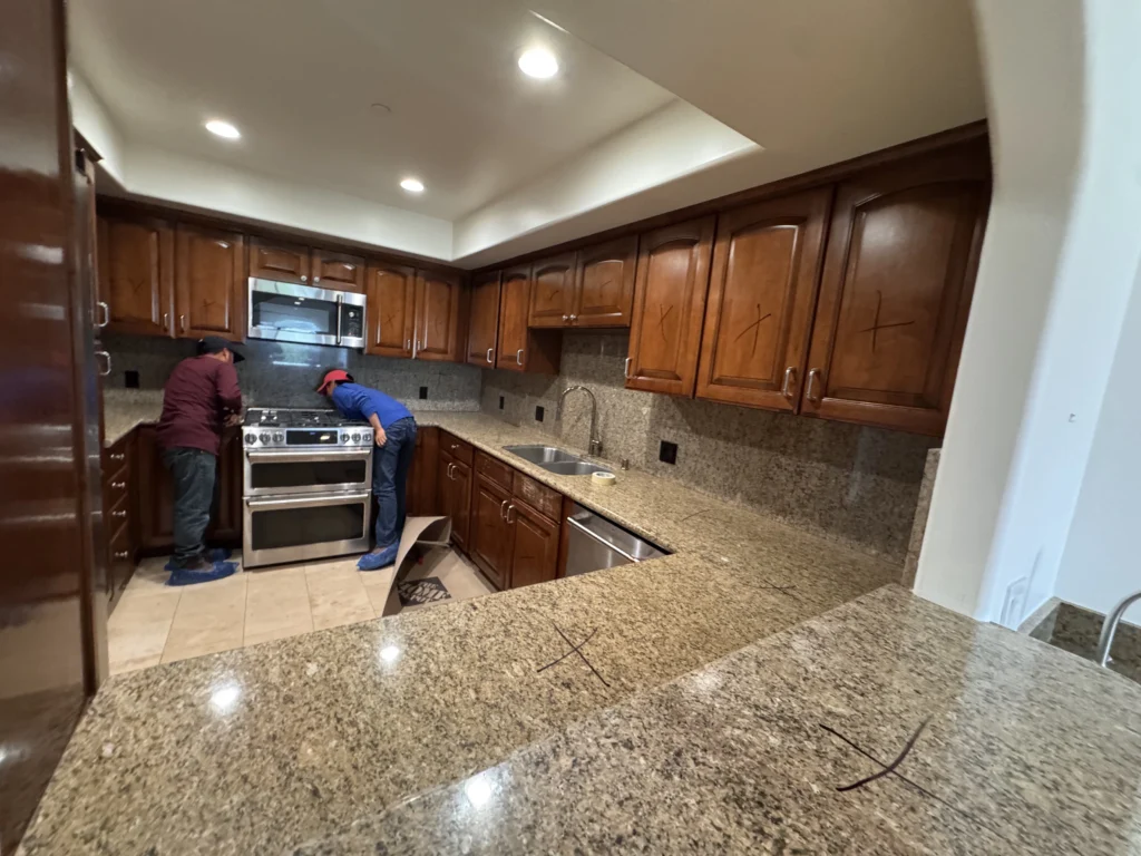 A wide shot of a kitchen with dark wood cabinets and granite countertops being worked on by contractors.