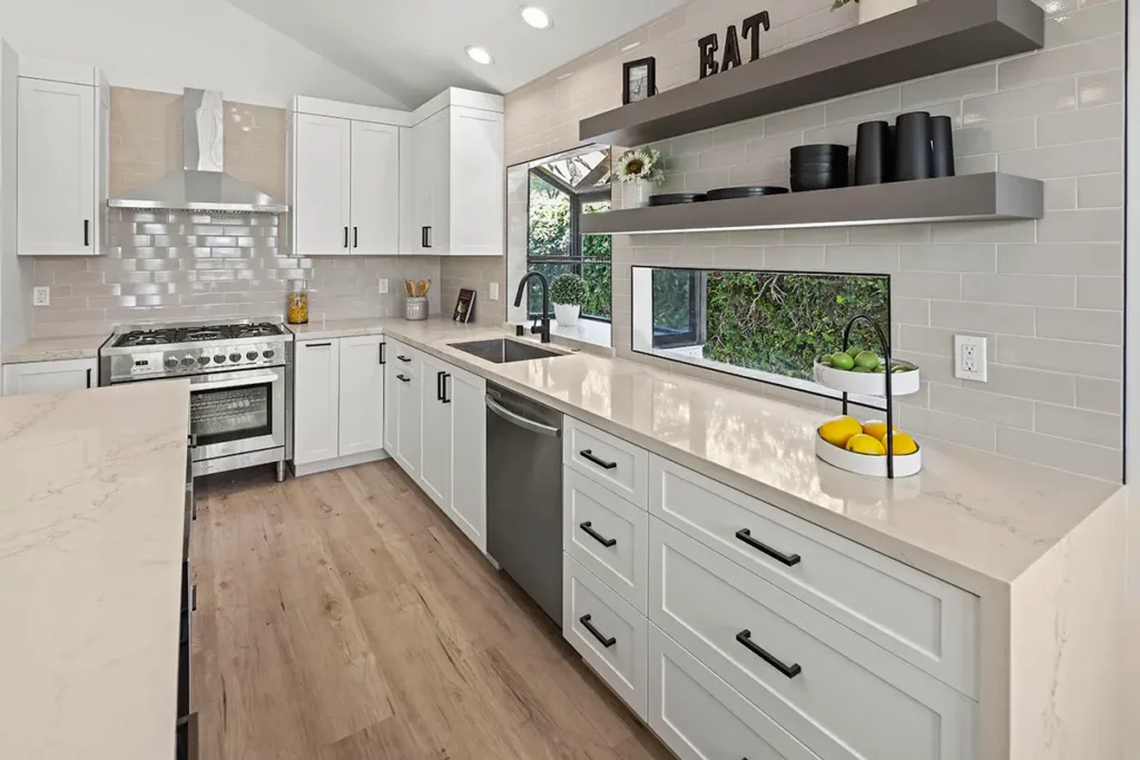 A view of a white kitchen with a tan countertop, stainless steel range, and two dark floating shelves on a tile backsplash.