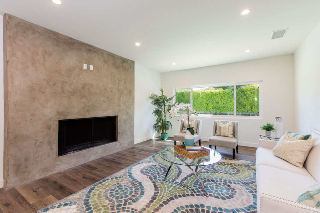 A bright, modern living room with hardwood floors, a large textured stone fireplace wall, and a colorful geometric area rug.