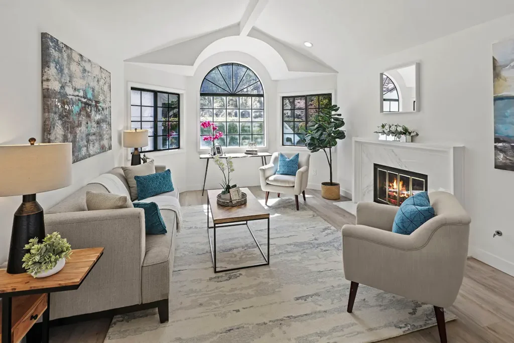 A formal living room featuring a white fireplace mantel, arched window, and two tan armchairs.