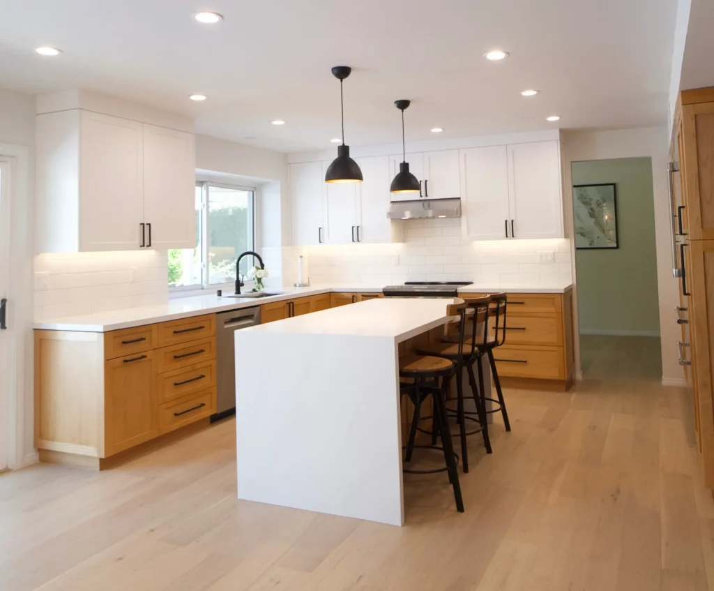 A wide view of a bright kitchen featuring a large white waterfall island, black pendant lights, and hardwood floors.