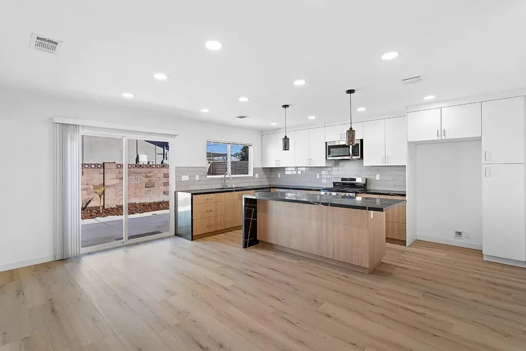 A wide view of a modern kitchen with a large island, wood-grain lower cabinets, and sliding glass doors leading outside.