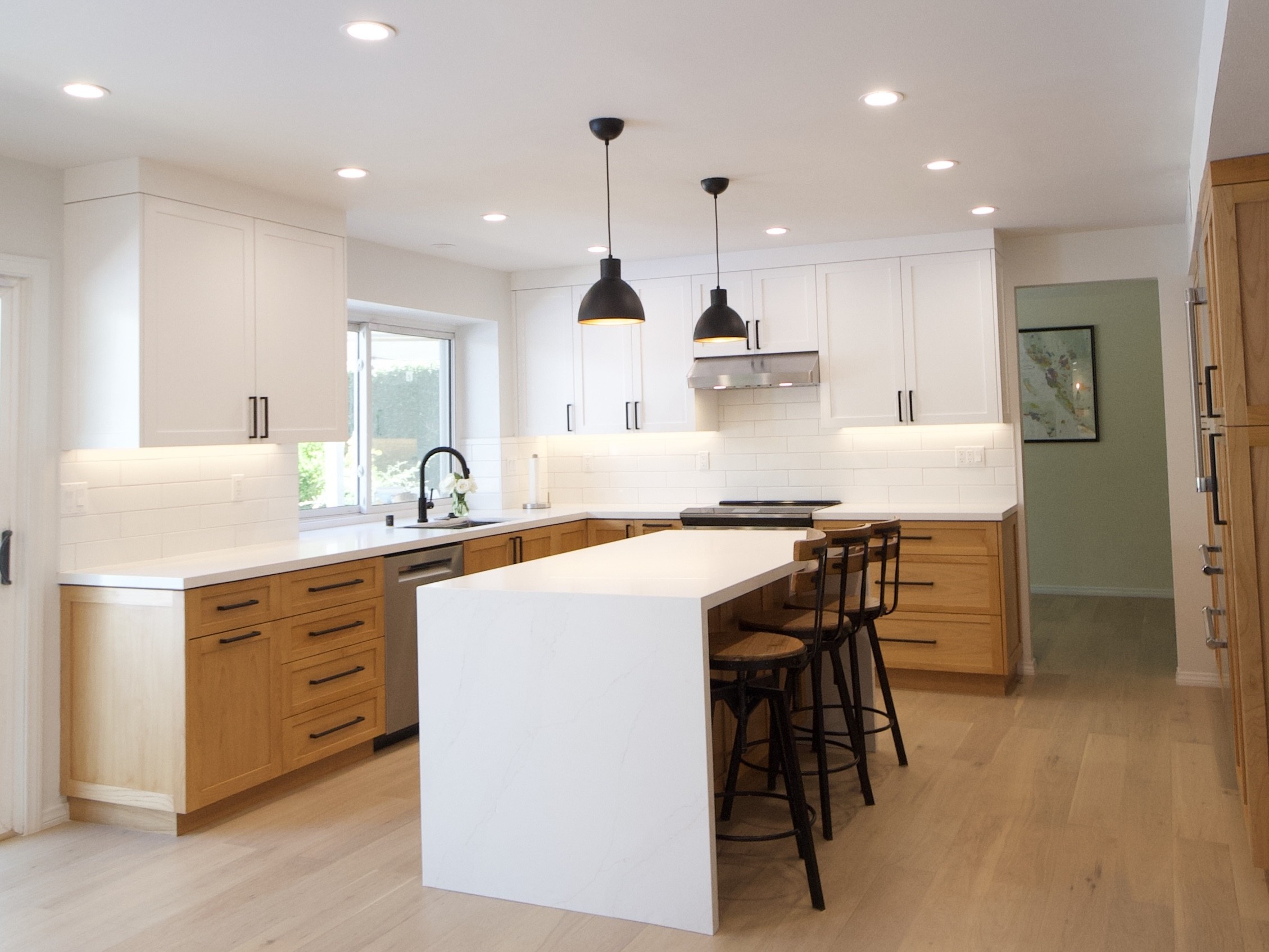 A wide shot of the kitchen showing the island with black barstools and the light hardwood flooring.