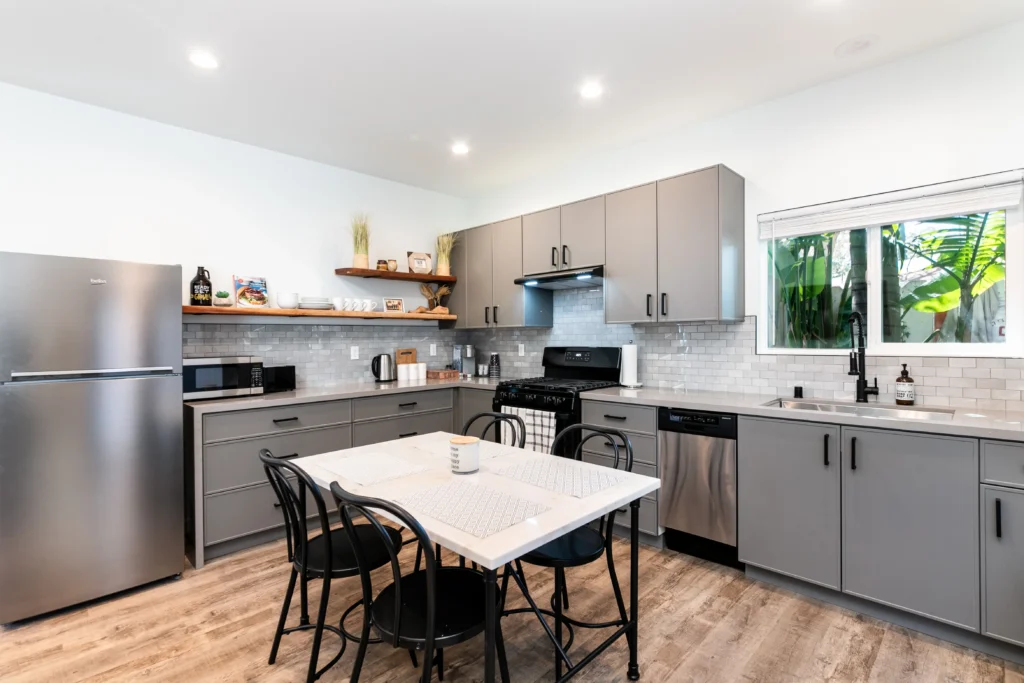 A wide interior view of an open-plan kitchen and dining space. It has modern grey cabinets, quartz countertops, a white subway tile backsplash, and stainless steel appliances. A small dark wood dining table with four chairs sits on the wood-look floor. Recessed lighting is in the white ceiling.