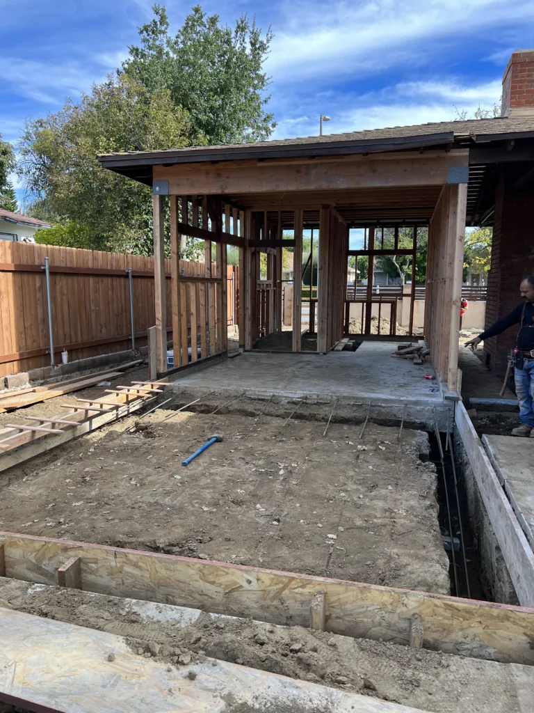 A backyard construction site featuring poured concrete slabs, retaining walls, and a framed patio cover structure.