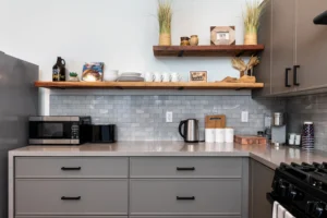 A focused shot of a modern L-shaped kitchen corner. It features grey shaker cabinets, a quartz countertop, and a white subway tile backsplash. Above the counter are two open natural wood shelves holding curated bottles and plants. Stainless steel appliances (refrigerator and range) are on the far left and right.
