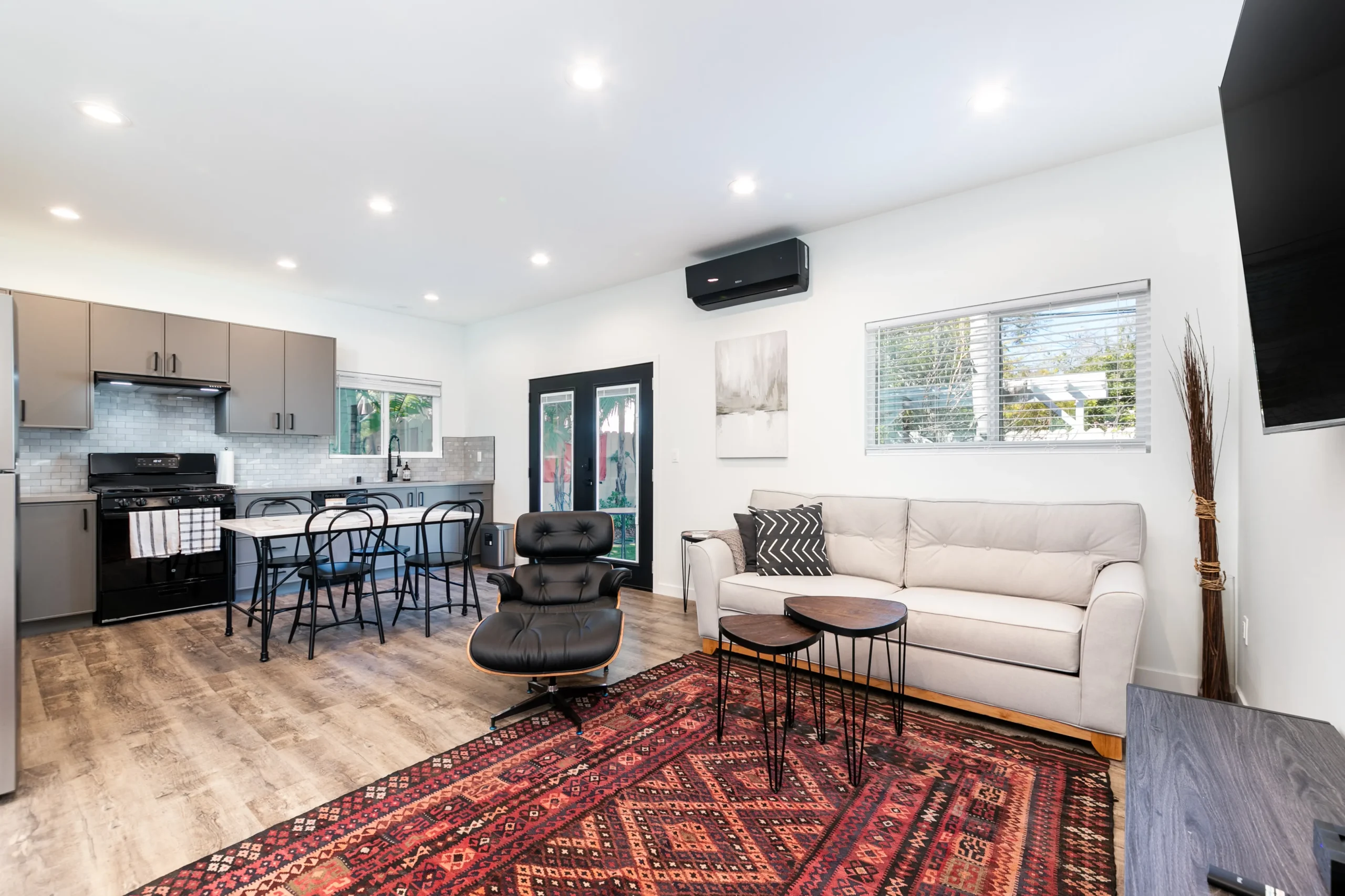 A wide-angle interior photograph taken from the kitchen corner. It shows the full length of the modern L-shaped kitchen with grey cabinets and stainless steel appliances, which opens up to a living room with a grey sofa, black lounge chair, and a large Persian rug. Multiple windows and recessed lighting illuminate the entire space.