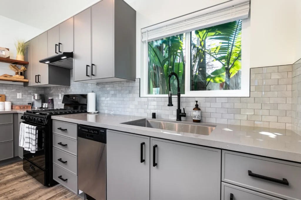 An interior view looking across a modern L-shaped kitchen. Grey cabinets and stainless steel appliances line the back wall. To the right, a large dark-framed glass door with matching windows opens directly onto an outdoor patio with plants and privacy screening.
