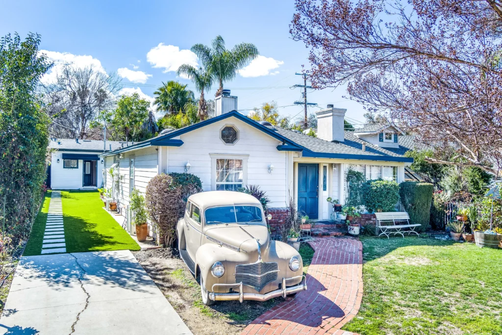 An aerial drone photograph of the front of a suburban property. A white single-story home with a blue door is visible. A unique, vintage brown vehicle is parked on a paved driveway in the foreground. Green artificial turf covers the yard. The ADU is partially visible behind the house.